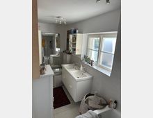 Kleines Badezimmer mit einem Doppelwaschbecken, weißen Badmöbeln und einem großen Fenster, das Tageslicht hereinlässt. Rechts im Bild steht ein geflochtener Wäschekorb neben einer Badewanne, und links sind ein Spiegel und ein Regal mit Hygieneartikeln zu sehen.