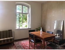 Ein schlichter, rustikaler Raum mit einem hölzernen Esstisch und vier Stühlen, einer gemusterten roten Fläche als Teppich, einem großen Fenster mit geöffnetem Flügel, sowie einer beigen Wand mit sichtbaren Spuren. Im Hintergrund stehen eine Zimmerpflanze und ein großer Spiegel, der auf den Boden gelehnt ist. Links befindet sich ein gestreiftes Sofa.