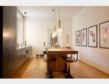 Moderne Küche mit hellen Wänden, einem Holzboden im Fischgrätmuster und einem Esstisch aus Holz mit Barhockern. Links befinden sich flache, dunkle Einbauschränke und eine Arbeitsplatte mit Spüle, während an der rechten Wand gerahmte Poster hängen.