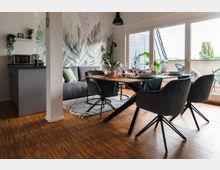 Modernes Wohnzimmer mit Essbereich, grauem Sofa, Holzfußboden und hellem Tageslicht durch große Fenster. Der Essbereich besteht aus einem Holztisch mit vier gepolsterten Stühlen, daneben eine kleine offene Küche; Pflanzen und Wanddekor ergänzen die gemütliche Atmosphäre.