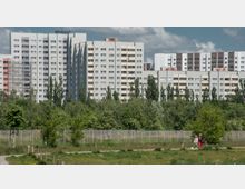 Wohnsiedlung mit hohen Plattenbauten im Hintergrund, umgeben von Bäumen und Grünflächen. Im Vordergrund ein umzäuntes Areal und zwei Personen auf einem Weg, begleitet von einem kleinen Hund.