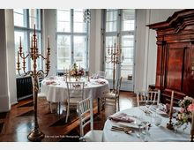 Eleganter Speisesaal mit hohen, weiß gerahmten Fenstern und Holzboden, darin runde, weiß gedeckte Tische mit Blumenarrangements, Gläsern und Besteck. Mehrarmige goldene Kerzenständer und ein großer dunkler Holzschrank prägen den Raum.