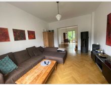 Wohnzimmer in einer Wohnung mit braunem Ecksofa, Holztisch und Parkettboden. Im Hintergrund ein Esstisch vor einer großen Balkontür, ein Kaminofen steht rechts an der Wand; moderne Kunstwerke hängen an den weißen Wänden.