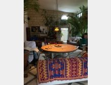 Gemütlicher Wohnraum mit einem runden Holztisch in der Mitte, umgeben von Sofas, die mit farbigen Decken im Ethnomuster bedeckt sind. Im Hintergrund befinden sich große grüne Pflanzen, eine Steinwand mit rundem Fenster sowie Bücherregale und dekorative Elemente.