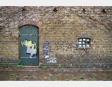 Eine Backsteinwand mit einer grünen Holztür und einem kleinen vergitterten Fenster. Die Wand ist mit Graffiti und Ranken verziert, der Boden besteht aus unregelmäßigem Kopfsteinpflaster.