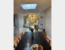 Ein modernes Esszimmer mit einem dunklen Holztisch, auf dem eine Vase mit orangefarbenen Tulpen und ein Teller mit Snacks steht. Darüber hängt eine große, kugelförmige Pendelleuchte aus Glas, und im Hintergrund ist ein heller Wohnbereich mit Sesseln und einem Bücherregal sichtbar. Ein Oberlicht im Dach lässt zusätzlich Tageslicht herein.