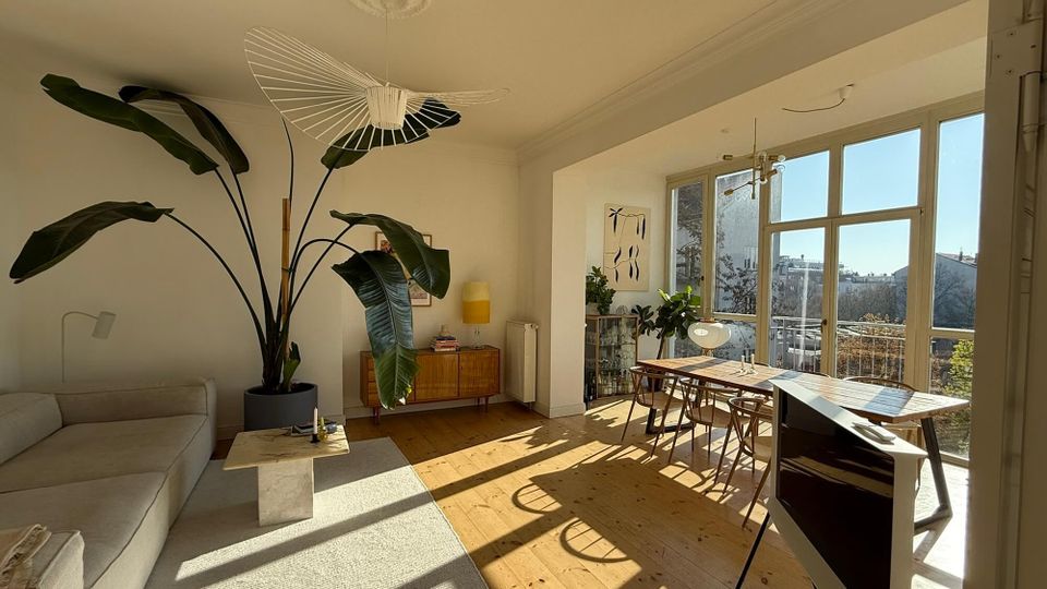 Bright open-plan living and dining room with light wood floors, a sofa seating area, large potted plant, and minimalist pendant lights. Floor-to-ceiling windows and a balcony door fill the space with sunlight, with a dining table and chairs set near the glass.