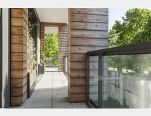 Moderner Balkonbereich mit Holzverkleidung an den Wänden, bodentiefen Glasgeländern sowie mit hellen Steinfliesen. Im Hintergrund sind grüne Bäume und ein parkähnlicher Außenbereich sichtbar.