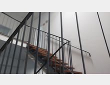 Eine moderne Innentreppe aus Holzstufen und einem schwarzen Metallgeländer, umgeben von einer weißen Wand und abgehängter Decke.