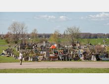 Eine Gemeinschaftsgartenanlage mit selbstgebauten Pflanzbeeten, Holzstrukturen und Dekorationen, umgeben von Wiesen und einzelnen Bäumen. Im Hintergrund ist eine weite, grüne Fläche sowie eine ziegelfarbene Gebäudereihe sichtbar.