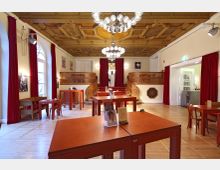 Innenraum eines historischen Cafés oder Veranstaltungsraums mit hölzernen Tischen und roten Vorhängen. Auffällig sind die dekorative Holzdecke, eine geschwungene Treppe mit Geländer auf beiden Seiten und ein Kronleuchter in der Mitte des Raumes.