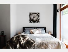 Schlafzimmer mit einem Doppelbett, dekoriert mit gemusterten Kissen und einer Tagesdecke mit goldenen Linien. An der weißen Wand hängt ein Bild, daneben ein großes Fenster mit dunklen Vorhängen und ein Nachttisch mit einer kleinen Lampe.