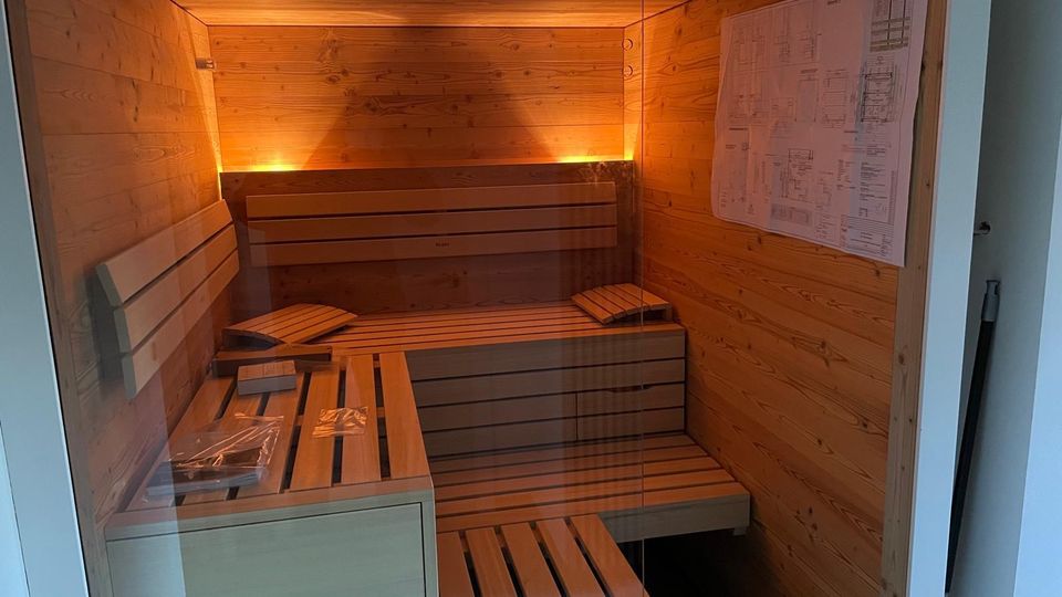 Blick in eine kleine Holzsauna mit Glasfront, umlaufenden Holzbänken in Stufenform und warmen indirekten Wandlichtern. An der rechten Wand hängt ein großer Plan, vorne steht eine niedrige Bank oder Ablage.