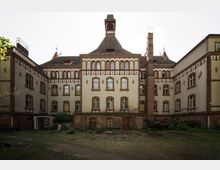 Historisches Backsteingebäude mit verzierten Fenstern und Klinkerfassaden-Elementen, das einen Innenhof umgibt. Das mehrstöckige Gebäude hat einen mittigen Turm mit Spitzdach und einen Schornstein, umgeben von überwachsenen Rasenflächen.