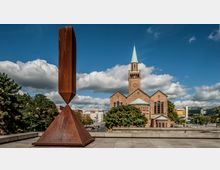 Eine historische Backsteinkirche mit grünem Kupferdach und Turm, umgeben von Bäumen und moderner Architektur im Hintergrund. Im Vordergrund steht eine große, rostfarbene Metallskulptur auf einem betonierten Platz unter einem blauen Himmel mit Wolken.