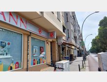 Straßenszene mit einer Hausfassade, die Geschäfte im Erdgeschoss zeigt, darunter eine Drogeriekette mit großen Schaufenstern und Werbeplakaten. Daneben befinden sich weitere kleine Läden und Cafés; am Bürgersteig stehen Fahrräder, ein E-Scooter und ein Stromkasten, während die Straße mit Bäumen gesäumt ist.