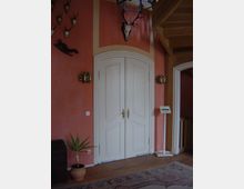 Innenraum mit rot-orange gestrichener Wand, einer weißen Doppeltür mit goldenen Griffen und Jagdtrophäen (Schädel und Geweihe) darüber. Links stehen eine Topfpflanze und eine Holzkiste, rechts befindet sich ein Treppenaufgang aus Holz, sowie ein Schild mit der Aufschrift „WC / Garderobe.“ Ein Teppich liegt auf dem Holzfußboden.