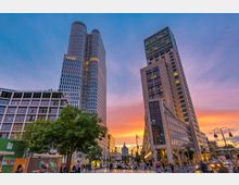 Städtische Szenerie in der Abenddämmerung mit zwei modernen Hochhäusern, darunter der Weiße Turm mit seiner charakteristischen abgerundeten Fassade. Im Vordergrund befinden sich Bäume, Passanten und Straßenelemente, während der Himmel in orange-rosa Tönen leuchtet.