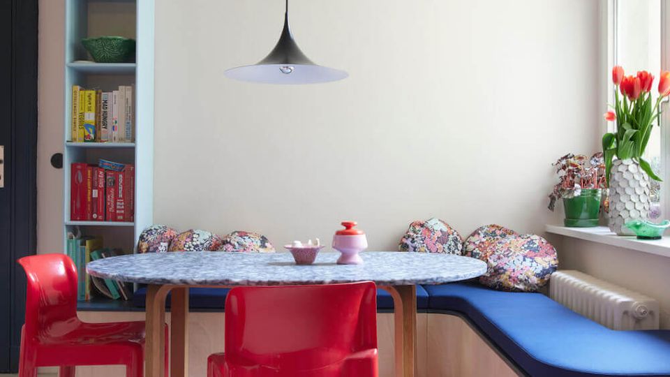 Bright dining nook with a round table and red plastic chairs, an L-shaped built-in bench with blue cushions and patterned pillows, and a black pendant light overhead. A tall narrow bookshelf stands beside a dark door, and large windows with flower vases sit above a radiator.