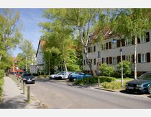 Ruhige Wohnstraße mit mehrstöckigen Häusern im traditionellen Baustil, weißen Fassaden und braunen Fensterläden. Am Straßenrand stehen Autos geparkt, und beidseitig der Straße wachsen Birken und weitere grüne Bäume.