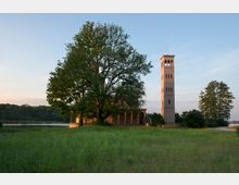 Historische Backsteinkirche mit freistehendem Glockenturm im romanischen Stil, umgeben von grünen Bäumen und einer Wiese; im Hintergrund ein See und bewaldete Hügel unter klarem Himmel.