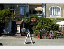 Straßenansicht eines gemütlichen Cafés mit dem Namen „Übersee Café · Bar“, umgeben von grüner Rankbepflanzung. Vor dem Gebäude stehen ein rotes Fahrrad, mehrere Tafeln mit Angeboten, sowie ein freistehendes Hinweisschild; daneben sind Sitzmöglichkeiten im Außenbereich erkennbar.