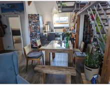 Offener Wohnraum in einem Dachgeschoss mit sichtbaren Holzträgern, einem Essbereich mit rustikalem Holztisch und Stühlen, einer Küche mit Dachfenster und modernen Möbeln sowie einer Metalltreppe, die zu einem Zwischengeschoss führt. Zimmerpflanzen und persönliche Dekorationen verleihen dem Raum eine wohnliche Atmosphäre.