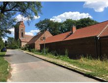 Straße in einem ländlichen Dorf mit einer Kirche aus Backstein im Hintergrund, deren Turm mit spitzem Dach hervorragt. Rechts stehen flache Gebäude mit roten Ziegeldächern, umgeben von Bäumen und Grünflächen unter einem blauen Himmel mit vereinzelten Wolken.