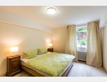 Ein schlichtes Schlafzimmer mit einem Doppelbett, bedeckt mit grüner Bettwäsche mit Blumenmuster. Zwei Nachttische mit Lampen stehen an den Seiten des Bettes, und ein Fenster mit hellen Vorhängen lässt Tageslicht in den Raum.