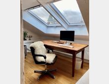 Ein Dachgeschoss-Arbeitszimmer mit einem Holzboden, einem Schreibtisch aus Holz, einem modernen Computer und einem schwarzen Bürostuhl mit weißem Fellbezug. Ein großes Dachfenster bietet Blick auf ein grünes Kirchturmdach und den Himmel.