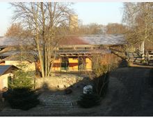 Ein ländliches Gebäude mit Holz- und Steinarchitektur, umgeben von Bäumen und befestigten Wegen. Im Vordergrund befindet sich ein Hof mit gepflasterten Flächen und vereinzelten Tannenbäumen.