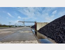 Industriegebiet mit einem großen Kohlelager rechts, waagerechte Kräne im Hintergrund und einer Betonfläche mit Wasserpfützen im Vordergrund. Ein Bogen einer Brücke und Strommasten sind in der Ferne sichtbar.