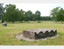 Park mit offenen Grünflächen und vereinzelten Bäumen; im Vordergrund eine wellenförmige Bankkonstruktion aus Holz und Beton mit Graffiti, daneben ein bunter Müllcontainer.