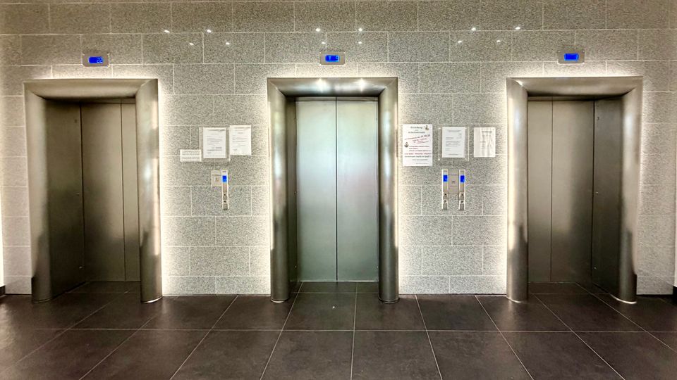 Drei moderne Aufzugstüren in einer Wand aus grauen Fliesen, jeweils mit blauen digitalen Stockwerksanzeigen und Tastenfeldern. Der Boden besteht aus dunklen Fliesen, und an der Wand befinden sich Aushänge und Informationen.