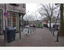 Pflastersteinstraße mit einem Brillengeschäft auf der linken Seite und verschiedenen Marktständen im Hintergrund. Karge Bäume säumen den Platz, der von Fußgängern und vereinzelten Fahrzeugen genutzt wird.