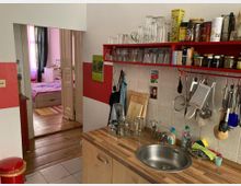 Kleine Küche mit einer Arbeitsplatte aus Holz, Spülbecken und Hängeregal mit Gläsern, Gewürzen und Utensilien. Im Hintergrund führt eine offene Tür in ein Schlafzimmer mit Holzboden und einem Bett.