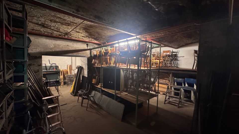 Ein Kellerraum mit gewölbter Decke und rohen Betonwänden, der als Lager genutzt wird. Verschiedene gestapelte Stühle, Leitern und Metallregale sind sichtbar, teilweise auf erhöhten Plattformen gelagert.