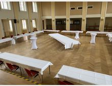 Großer, heller Saal mit Parkettboden, hohen Fenstern und cremefarbenen Wänden. Die Tische sind mit weißen Tischdecken gedeckt und in einer U-Form angeordnet, ergänzt durch einige Stehtische mit weißen Hussen.