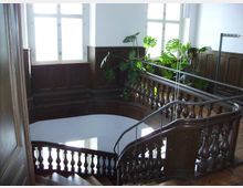 Holztreppenhaus mit kunstvoll geschnitztem Geländer und einer Podestfläche, umgeben von dunkler Holzvertäfelung. Große Fenster lassen Tageslicht herein, und grüne Zimmerpflanzen schmücken den Bereich.