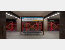 U-Bahn-Station Jungfernheide der Linie U7 mit einem zentralen Sitzbereich, gelben Hinweisschildern und roten Gitterabsperrungen im Hintergrund. Die Beleuchtung ist schlicht und die Wände im Hintergrund zeigen eine bunte Wandgestaltung.