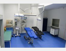 Moderner Operationssaal mit blauen Böden, zwei Untersuchungsliegen, medizinischen Geräten und Hängelampen. An den Wänden befinden sich Regale, ein Schrank und ein Fenster mit Blick in einen angrenzenden Raum.