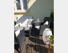Gemütliche Terrasse mit Holzboden, schwarzen Rattanmöbeln und weißen Kissen, dekoriert mit einer Laterne, Blumen und einem kleinen Tisch mit Glasgefäßen. Im Hintergrund ist eine helle Hauswand mit einem großen Fenster zu sehen.
