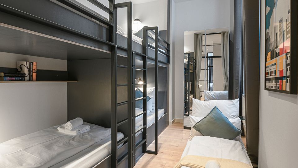 Schmales, modern eingerichtetes Schlafzimmer mit schwarzen Metall-Etagenbetten auf der linken Seite und einem Einzelbett am Fenster rechts. Helle Holzböden, weiße Wände, Einbauspots an der Decke sowie ein Spiegel und ein Vorhang am Ende des Raums.