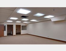 Leerer Büroraum mit beigefarbenem Teppichboden, weißen Wänden und abgehängter Decke mit eingebauten quadratischen Deckenleuchten; an der Wand sind eine geschlossene Tür, zwei offene Türrahmen und ein kleines Fenster eingebaut.