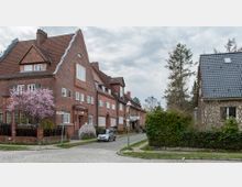Straßenszene mit roten Backsteinhäusern im traditionellen Baustil, einer blühenden rosa Baumkrone im Vorgarten und einem kleinen, gepflasterten Gehweg. Im Vordergrund ein Straßenschild mit der Aufschrift „Falkenried“ und ein geparktes Auto, rechts eine Hecke und ein weiteres Haus mit grauem Dach.