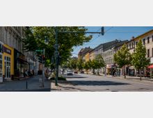 Straßenszene in einer Stadt mit mehrstöckigen Wohn- und Geschäftsgebäuden auf beiden Seiten, darunter Geschäfte und Cafés. Entlang der Straße stehen Bäume, und es gibt Fußgänger sowie fahrenden Verkehr; eine Ampel und Straßenschilder sind ebenfalls sichtbar.