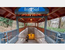 Eingang zur U-Bahnstation Britz-Süd mit einem überdachten Holzvordach, Metallgeländern und Treppen, die in einen unterirdischen Bereich führen. Im Hintergrund sind Bäume und Sträucher zu sehen.