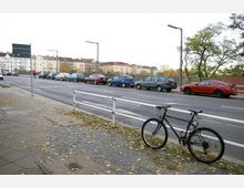 Straßenansicht mit einem Fahrrad, das an einem weißen Geländer auf einem gepflasterten Gehweg lehnt. Im Hintergrund parkende Autos entlang einer Straße, Wohnhäuser mit roten Dächern und herbstliche Bäume.