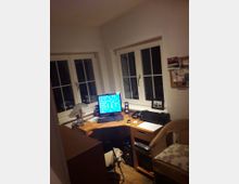 Kleines Arbeitszimmer mit zwei Fenstern, einem Holzschreibtisch in der Ecke und einem PC mit Monitor, Tastatur und Lautsprechern. Rechts steht ein Korbstuhl, an der Wand gegenüber hängt ein Pinnbrett mit Notizen.