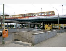 Straßenszene mit erhöhtem S-Bahngleis, auf dem ein Zug fährt, und einer darunter liegenden Bushaltestelle mit Glasüberdachung. Im Vordergrund befindet sich ein Treppenabgang zur U-Bahn, daneben ein orangefarbener Mülleimer und ein parkender DHL-Transporter.